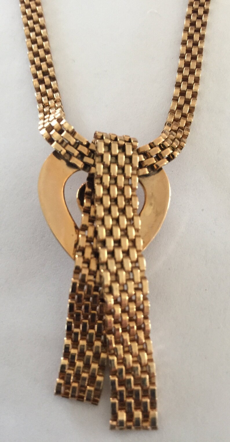 May include: A gold chain necklace with a heart-shaped pendant. The chain is made of small, square links. The pendant is a heart with a cutout in the center. The chain is tied in a bow at the bottom of the pendant.