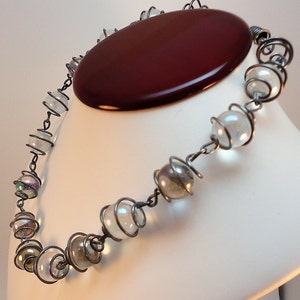 May include: A silver wire necklace with iridescent glass beads. The necklace is on a white mannequin bust.