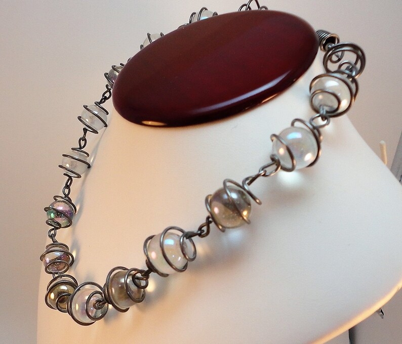 May include: A silver wire necklace with iridescent glass beads. The necklace is on a white mannequin bust.