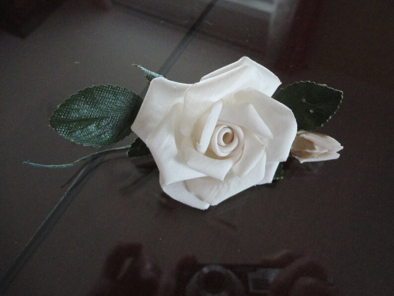 Puede incluir: Una sola rosa blanca de tela con hojas verdes. La rosa es un dise&ntilde;o simple y elegante.