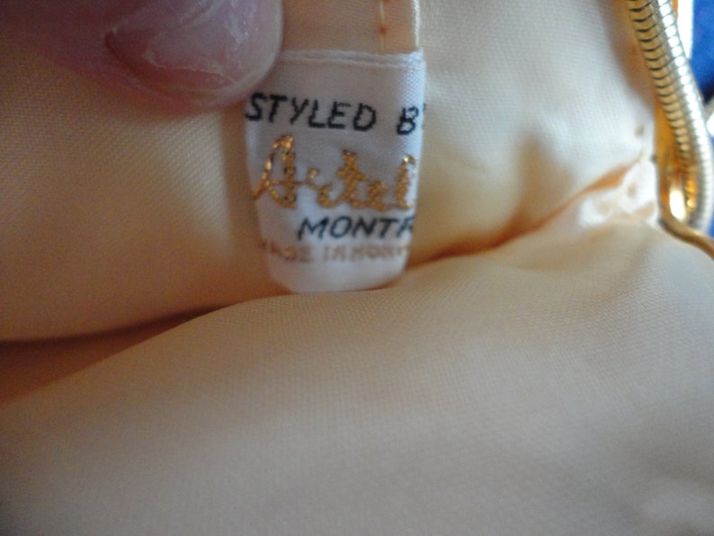May include: A white satin clutch bag with a gold chain strap. The bag has a label that reads "Styled by Artel, Mont-R, Made in Hong Kong".