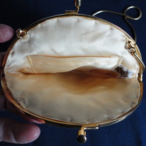 May include: A gold-toned metal purse with a round, cream-colored satin interior. The purse has a clasp closure and a chain strap.