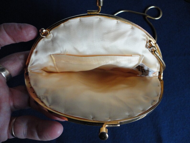 May include: A gold-toned metal purse with a round, cream-colored satin interior. The purse has a clasp closure and a chain strap.