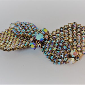 May include: A gold-toned brooch with a swirling design made of iridescent rhinestones. The rhinestones are arranged in a pattern that resembles a wave or a ribbon.