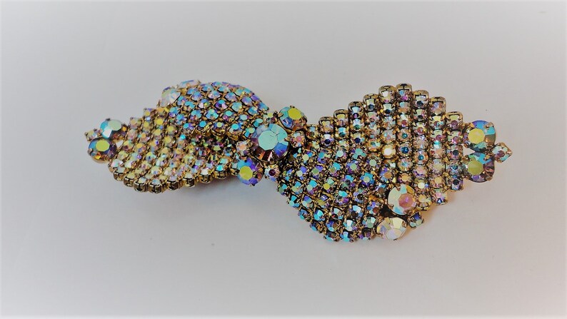 May include: A gold-toned brooch with a swirling design made of iridescent rhinestones. The rhinestones are arranged in a pattern that resembles a wave or a ribbon.