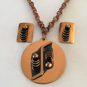 May include: A copper-toned jewelry set featuring a necklace and matching earrings. The necklace has a round pendant with a geometric design and a chain. The earrings are rectangular with a similar design. The set has a vintage aesthetic.