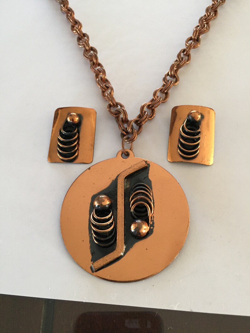 May include: A copper-toned jewelry set featuring a necklace and matching earrings. The necklace has a round pendant with a geometric design and a chain. The earrings are rectangular with a similar design. The set has a vintage aesthetic.