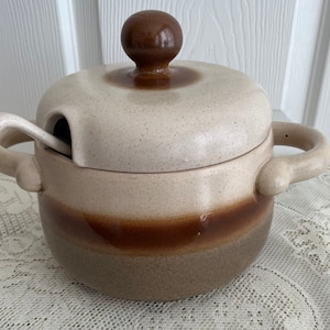 Peut inclure: Une soupière en céramique brune et blanche avec un couvercle et une louche assortie. La soupière a une bande brune autour du milieu et un bouton brun sur le couvercle.
