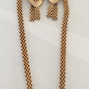 May include: A gold tone necklace and earrings set with a heart pendant and dangling chains. The necklace and earrings are made of a woven chain pattern.