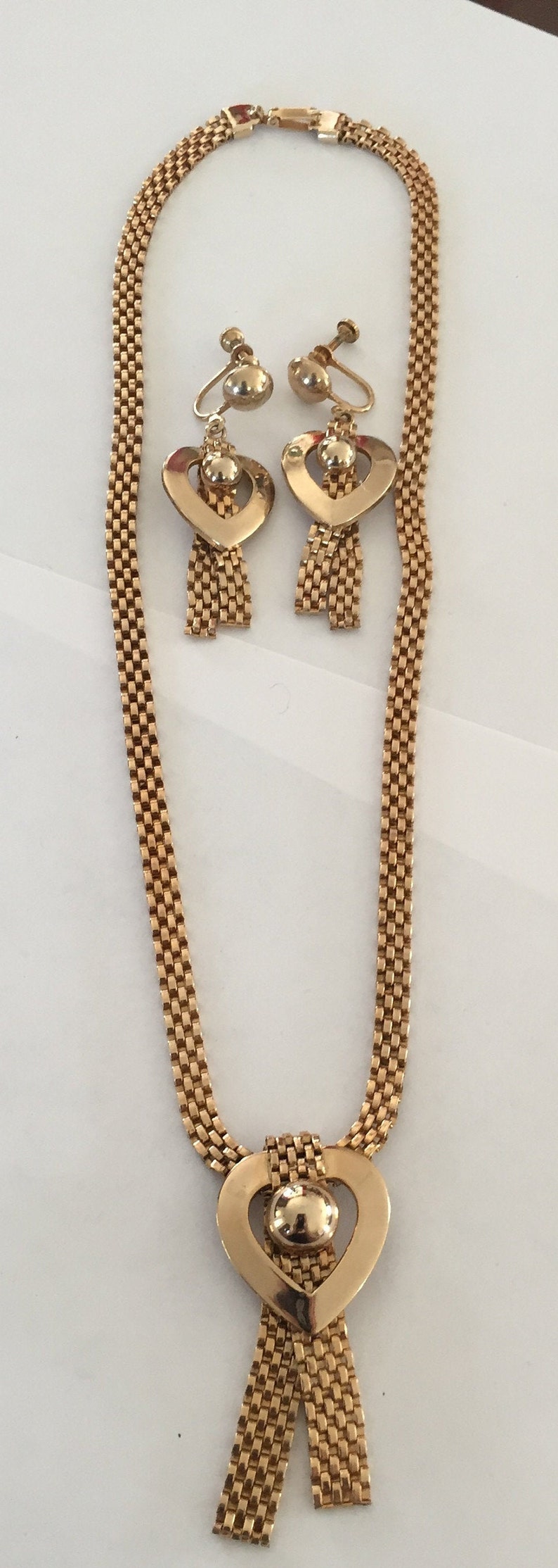 May include: A gold tone necklace and earrings set with a heart pendant and dangling chains. The necklace and earrings are made of a woven chain pattern.