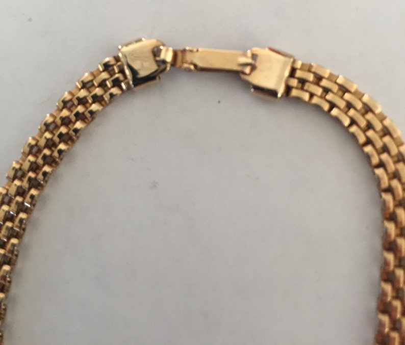 May include: A gold chain necklace with a box clasp closure. The chain is made of multiple rows of small, linked squares.