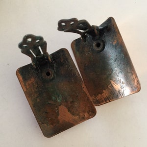 May include: A pair of vintage clip-on earrings. Each earring is a rectangular shape with rounded corners, featuring a copper-colored finish with areas of verdigris. The clip mechanism is a decorative, ornate design.