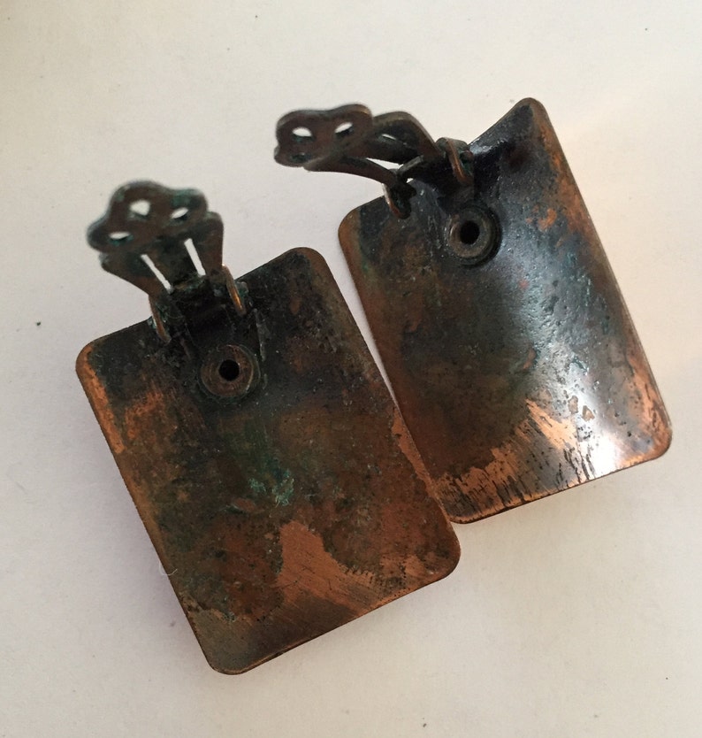 May include: A pair of vintage clip-on earrings. Each earring is a rectangular shape with rounded corners, featuring a copper-colored finish with areas of verdigris. The clip mechanism is a decorative, ornate design.