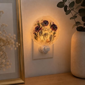 Può includere: Una luce notturna rotonda con un design floreale. La luce presenta una parte anteriore trasparente e strutturata con fiori secchi incorporati in tonalità di viola, giallo e verde. La luce notturna è collegata a una presa a muro bianca.