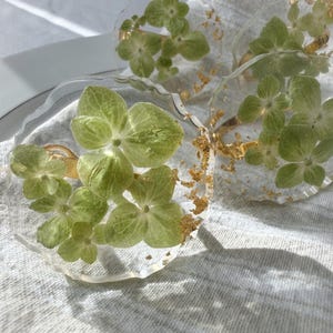 May include: Clear resin coasters with embedded green hydrangea flowers and gold leaf accents. The coasters have a unique, organic shape and are set on a light gray linen surface. These decorative coasters add a touch of nature to any home decor.
