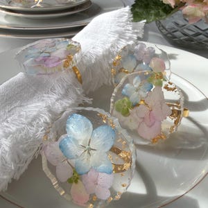 Könnte beinhalten: Transparente Serviettenringe aus Kunstharz mit eingebetteten blauen, rosa und grünen Hortensienblüten und Goldblattakzenten. Die Ringe sind auf goldfarbenen Metallbändern befestigt. Mehrere Ringe sind auf einem weißen Teller mit einer weißen Serviette abgebildet.