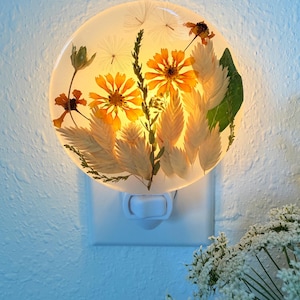 Könnte beinhalten: Ein rundes Nachtlicht mit getrockneten Blumen, die in klarem Harz eingeschlossen sind. Die Blumen umfassen orange und gelbe Blüten, grüne Blätter und federartige beige Akzente. Das Nachtlicht ist in eine weiße Steckdose gesteckt und strahlt ein warmes Licht aus.