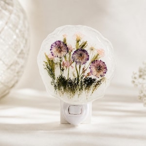 Pode incluir: Luz noturna decorativa com flores secas encapsuladas em resina transparente. As flores são roxas, rosas e amarelas, sobre um fundo branco. A luz noturna tem uma base branca e um interruptor liga/desliga.