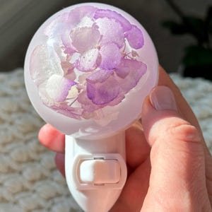 Op de afbeelding: Een witte nachtlamp met een bolvormige, heldere harsbol. De bol bevat gedroogde paarse en witte bloemen. De nachtlamp heeft een schakelaar en is ontworpen om in een stopcontact te worden gestoken.