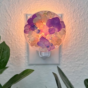 Op de afbeelding: Een decoratief nachtlampje met een helder, rond harsontwerp gevuld met gedroogde paarse, witte en gele bloemen. Het nachtlampje is aangesloten op een wit stopcontact en straalt een warme gloed uit.