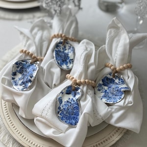 May include: Four white cloth napkins folded and secured with napkin rings made of natural wooden beads and blue and white floral patterned oyster shells with silver-toned edges, arranged on a white plate with a fringed border.