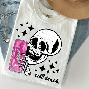 May include: White t-shirt featuring a black skull graphic holding a pink energy drink can. The can reads "Alani Energy Pink Slush" and the shirt has the text "till death." Black star shapes are scattered around the design.