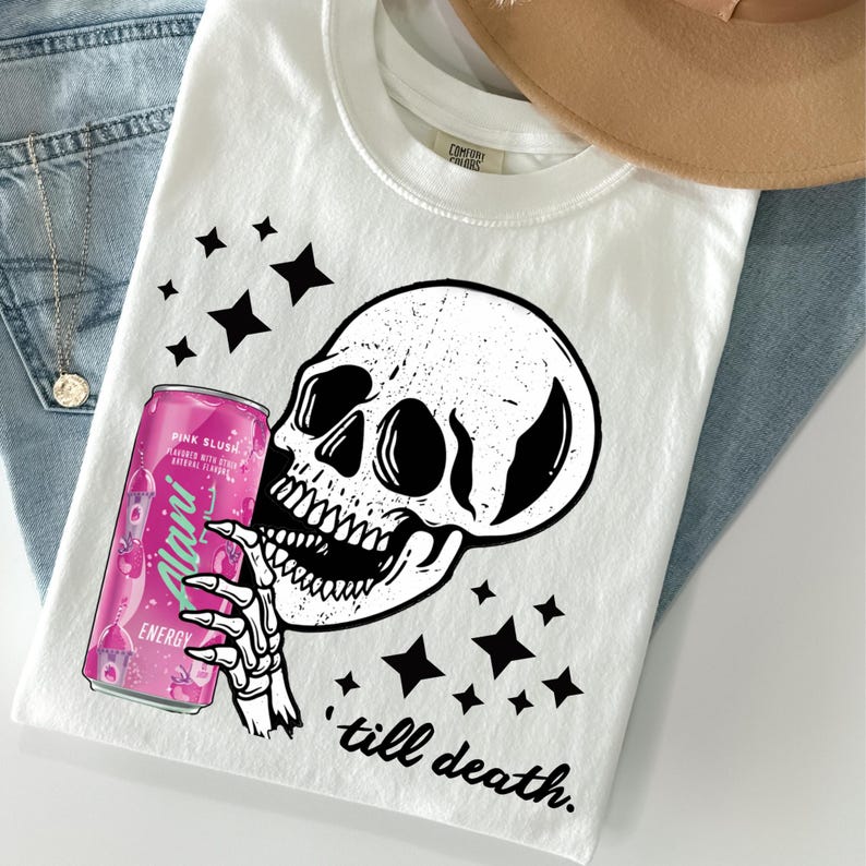 May include: White t-shirt featuring a black skull graphic holding a pink energy drink can. The can reads "Alani Energy Pink Slush" and the shirt has the text "till death." Black star shapes are scattered around the design.