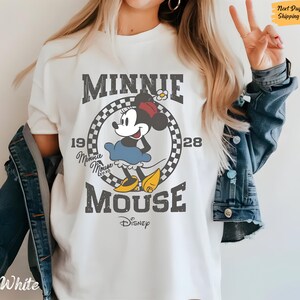 K&ouml;nnte beinhalten: Wei&szlig;es T-Shirt mit einem Vintage-Grafikdruck von Minnie Mouse. Die Grafik zeigt Minnie Mouse in einem roten Kleid und gelben Schuhen, mit dem Text "Minnie Mouse" und "Disney" in einer verwitterten Schriftart.