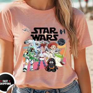 May include: Peach-colored t-shirt featuring a Star Wars graphic with cartoon characters. The design includes the Star Wars logo, a spaceship, and characters like Boba Fett, Woody, and Darth Vader. The shirt has a crew neck.