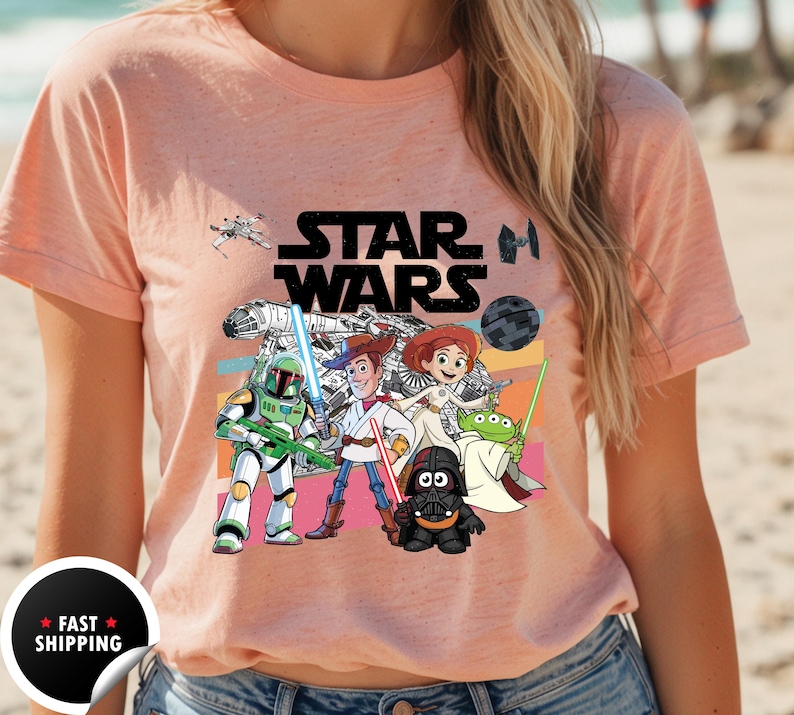 May include: Peach-colored t-shirt featuring a Star Wars graphic with cartoon characters. The design includes the Star Wars logo, a spaceship, and characters like Boba Fett, Woody, and Darth Vader. The shirt has a crew neck.