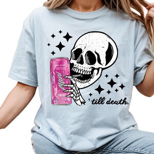 May include: Light blue t-shirt featuring a black and white skull graphic holding a pink energy drink can. The can reads "Pink Slush" and "Alani Energy". The phrase "till death" is printed below the skull.