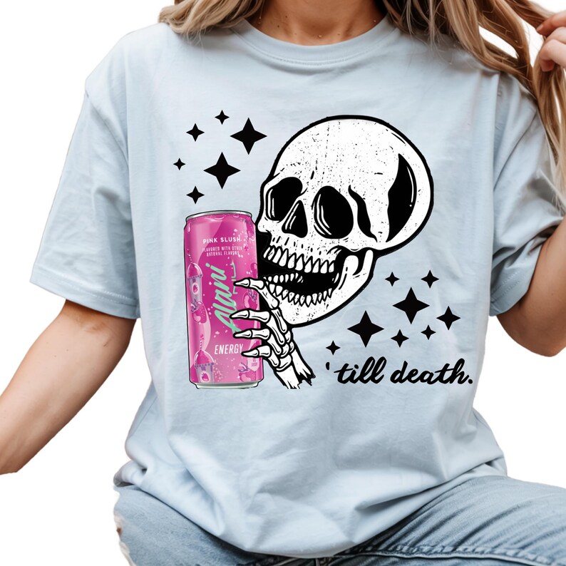 May include: Light blue t-shirt featuring a black and white skull graphic holding a pink energy drink can. The can reads "Pink Slush" and "Alani Energy". The phrase "till death" is printed below the skull.