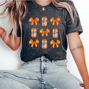 May include: A gray t-shirt with a repeated pattern of orange bows and orange and white cans of Fanta soda.