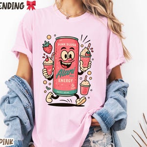 May include: Light pink t-shirt featuring a cartoon can of "Pink Slush" energy drink with a smiling face, holding two slushie cups. The shirt also has the words "TRENDING" and "LIGHT PINK" printed on it.