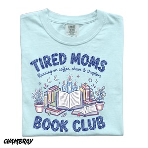 May include: Light blue t-shirt with the text "TIRED MOMS BOOK CLUB" in dark blue. The design includes illustrations of books, candles, plants, and stars. The shirt is made of Comfort Colors material.