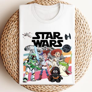 May include: White t-shirt featuring the words "STAR WARS" in large black letters. The shirt has a colorful graphic of Star Wars and Toy Story characters. The design includes a spaceship, a Death Star, and various characters.
