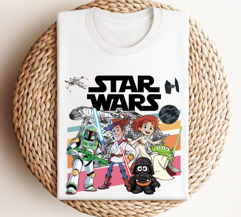 May include: White t-shirt featuring the words "STAR WARS" in large black letters. The shirt has a colorful graphic of Star Wars and Toy Story characters. The design includes a spaceship, a Death Star, and various characters.