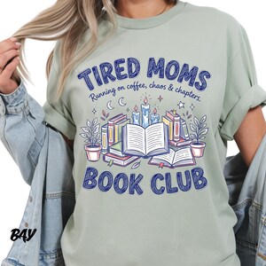 May include: Sage green t-shirt with the text "TIRED MOMS BOOK CLUB" in blue. The graphic includes books, candles, and plants. The phrase "Running on coffee, chaos & chapters" is also included.
