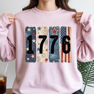 May include: A light pink sweatshirt featuring the number "1776" in black. The numbers are set against a backdrop of patriotic designs, including stars, fireworks, and the American flag. The sweatshirt is a comfortable, casual style.