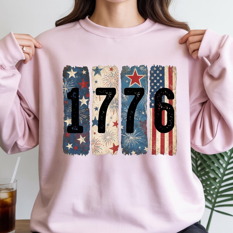 May include: A light pink sweatshirt featuring the number "1776" in black. The numbers are set against a backdrop of patriotic designs, including stars, fireworks, and the American flag. The sweatshirt is a comfortable, casual style.