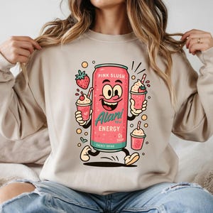 May include: A light beige sweatshirt featuring a cartoon design of a pink energy drink can labeled "Pink Slush" and "Alani Energy". The design includes cartoon strawberry drinks and strawberries.