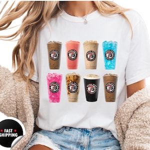 May include: White t-shirt featuring a variety of iced coffee drinks in clear plastic cups. Each cup has a different colored beverage and a logo with the text "Seven Brew". The shirt is worn by a person.