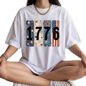May include: White t-shirt featuring the number "1776" in black, with each number set against a vertical rectangular design. The designs include fireworks, stars, and the American flag, creating a patriotic theme.