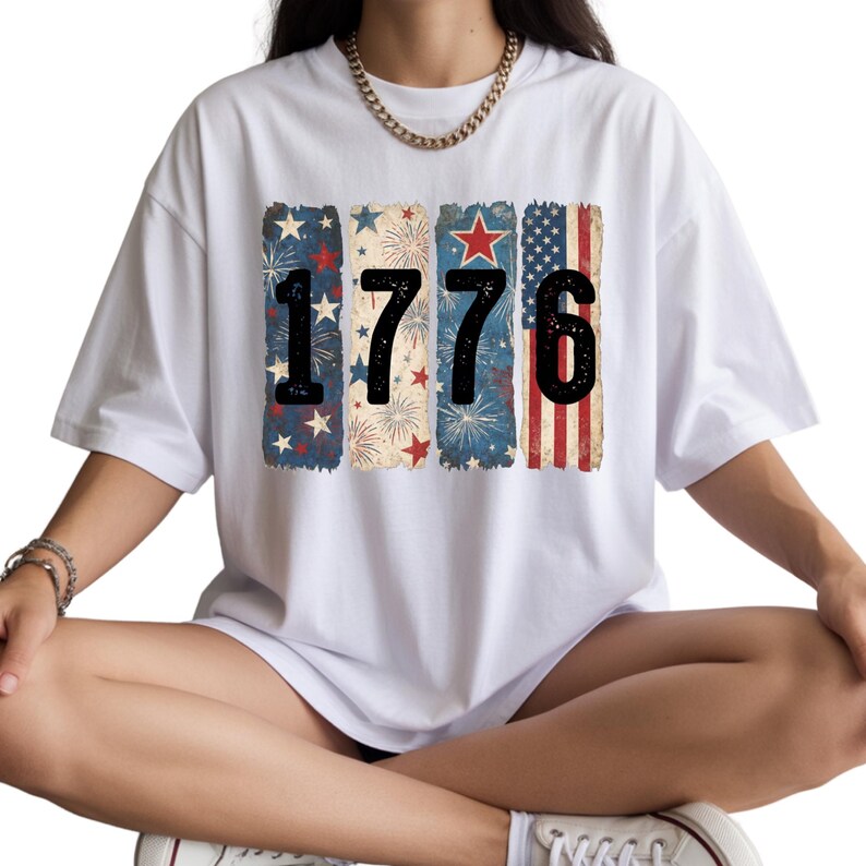 May include: White t-shirt featuring the number "1776" in black, with each number set against a vertical rectangular design. The designs include fireworks, stars, and the American flag, creating a patriotic theme.