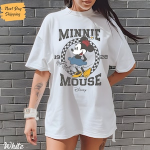 K&ouml;nnte beinhalten: Wei&szlig;es T-Shirt mit einem Vintage-Grafikdruck von Minnie Mouse. Die Grafik zeigt Minnie Mouse in einem blauen Kleid und roten Schuhen, mit dem Text "Minnie Mouse" und "1928" um sie herum. Der Text "Disney" befindet sich am unteren Rand der Grafik.
