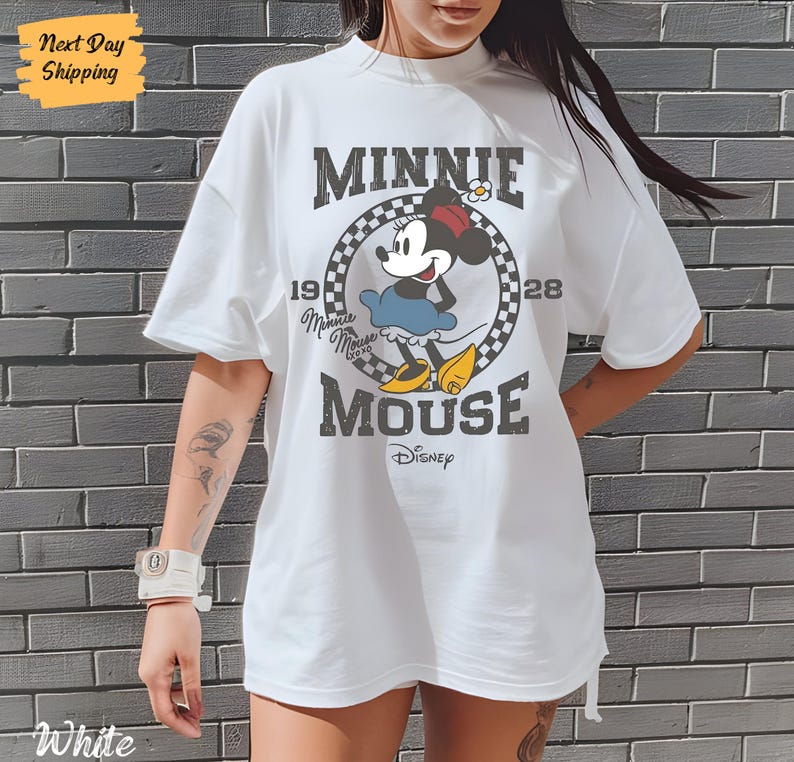 K&ouml;nnte beinhalten: Wei&szlig;es T-Shirt mit einem Vintage-Grafikdruck von Minnie Mouse. Die Grafik zeigt Minnie Mouse in einem blauen Kleid und roten Schuhen, mit dem Text "Minnie Mouse" und "1928" um sie herum. Der Text "Disney" befindet sich am unteren Rand der Grafik.