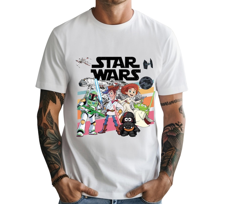 May include: White t-shirt featuring the Star Wars logo in black block letters above a colorful cartoon graphic of Star Wars characters. The graphic includes Boba Fett, Woody, Jessie, and other characters.