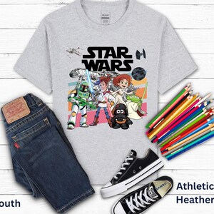 May include: A light gray youth t-shirt with a Star Wars graphic featuring characters and the Star Wars logo. The image includes a pair of dark blue jeans, black sneakers, and a bundle of colored pencils.