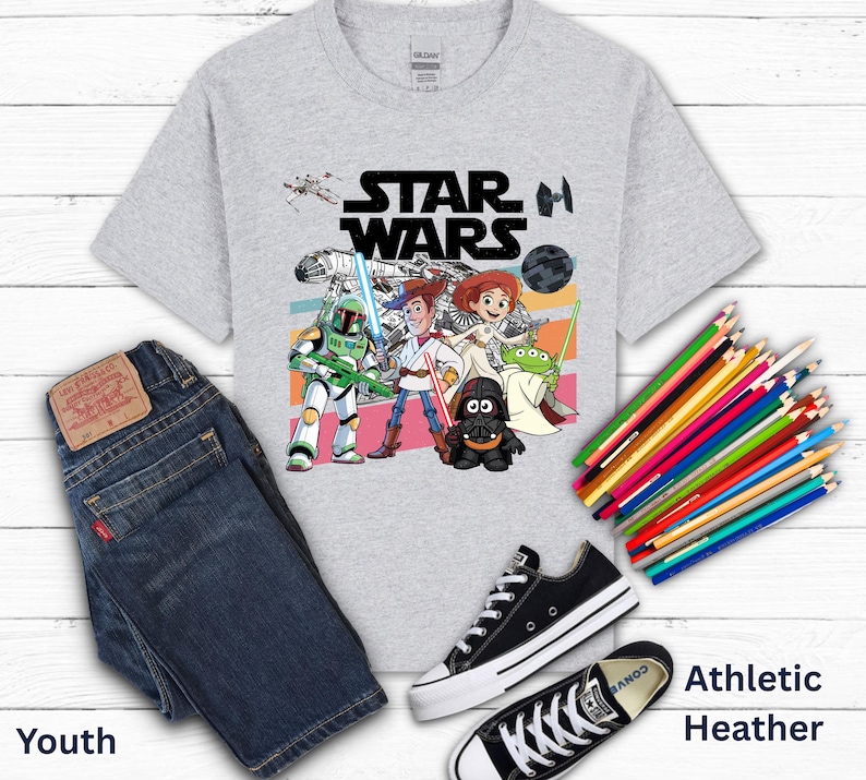May include: A light gray youth t-shirt with a Star Wars graphic featuring characters and the Star Wars logo. The image includes a pair of dark blue jeans, black sneakers, and a bundle of colored pencils.