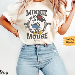 K&ouml;nnte beinhalten: Ein wei&szlig;es T-Shirt mit einem Vintage-Grafikdruck von Minnie Mouse. Die Grafik zeigt Minnie Mouse in einem roten Kleid und gelben Schuhen, mit dem Text "Minnie Mouse" und "Disney" darunter. Die Grafik ist von einem karierten Rand umgeben.